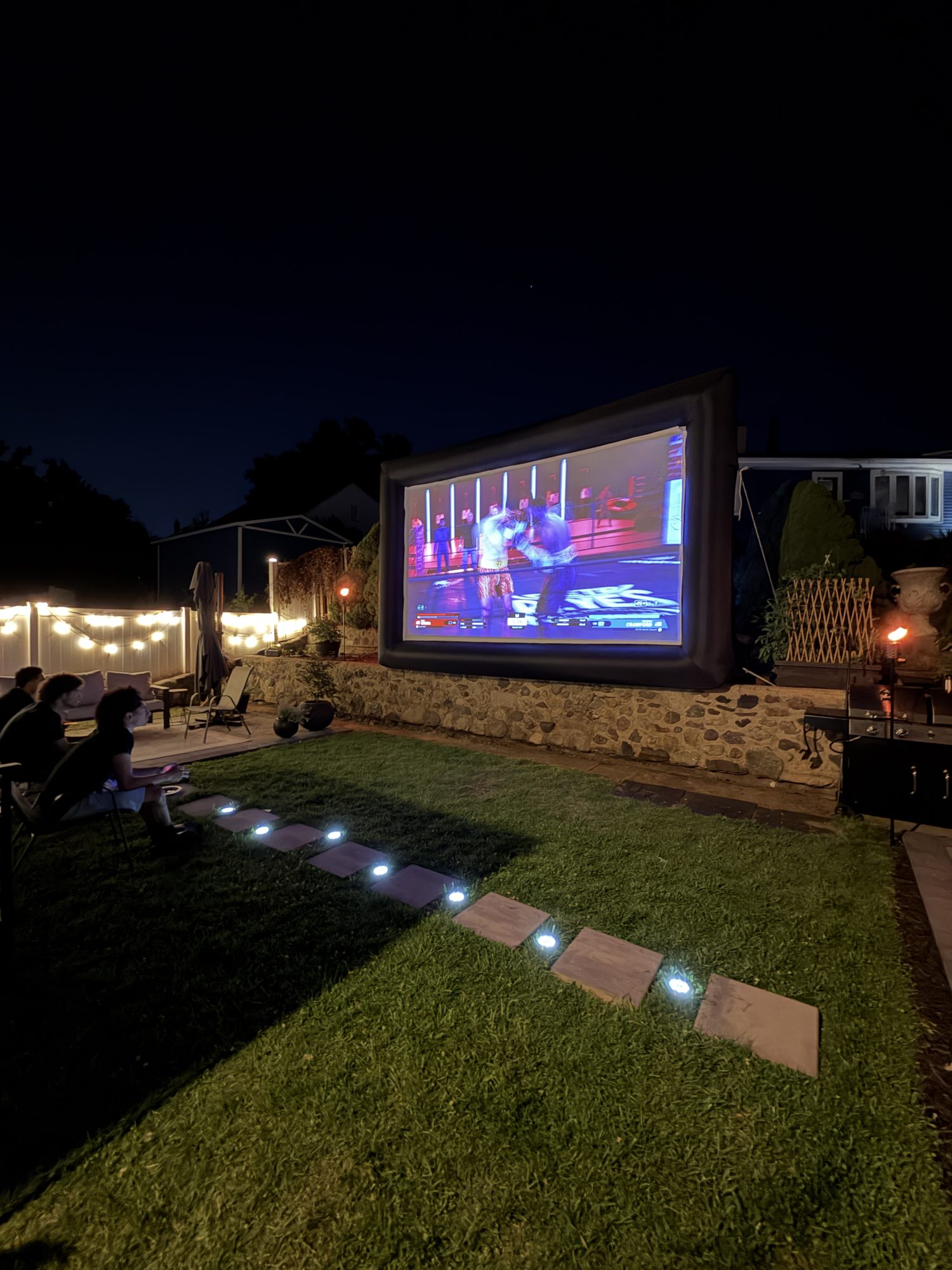 An outdoor movie playing on the Light Haus screen after dark, with string lights and a lit garden path